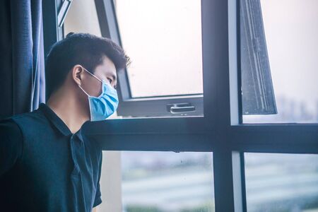 Quarantine self-isolation. Sad young man in a medical mask who looks out the window through the window. Infected man in medical mask on self-isolation looks at the street through the window of a houseの写真素材