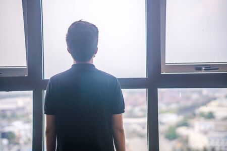 Quarantine self-isolation. Sad young man in a medical mask who looks out the window through the window. Infected man in medical mask on self-isolation looks at the street through the window of a houseの写真素材