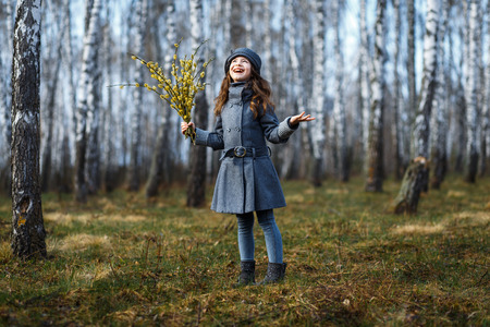 A girl in grey coat and a cute gray hat in the forest in early spring with a willow branch of twigs smelling twigs. A girl outside after rain at the sunsetの写真素材