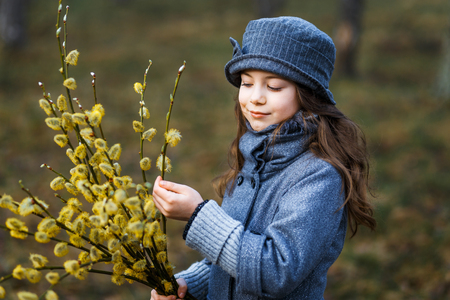 A girl in grey coat and a cute gray hat in the forest in early spring with a willow branch of twigs smelling twigs. A girl outside after rain at the sunsetの写真素材