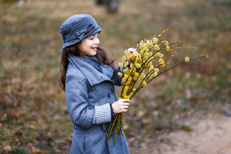A girl in grey coat and a cute gray hat in the forest in early spring with a willow branch of twigs smelling twigs. A girl outside after rain at the sunsetの写真素材