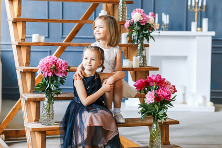 Two cute little girls with peonies. Two beautiful friends having fun, hugging and smilingの写真素材