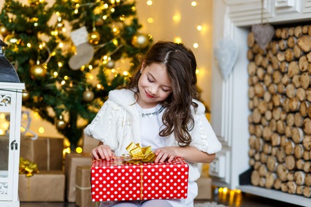 A brunette gilr in front of fur-tree and fireplace with candles and gifts. A surprised girl. A girl dreaming. New year's eve. Christmas eve. Cozy holiday at the fur-tree with lights and gold decor.の写真素材