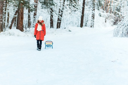 Beautiful teenage girl in a long red down jacket white hat and a scarf having fun outside in a wood with snow in winterの写真素材