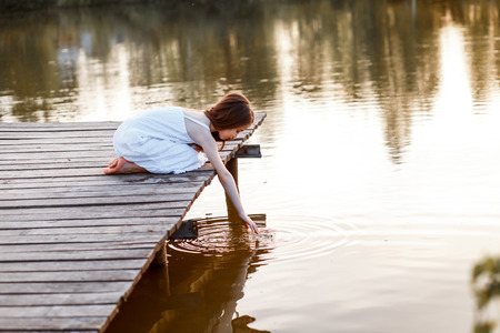 A beautiful girl with long brunette hair and blue eyes having fun on the pier near the water at the sunsetの写真素材