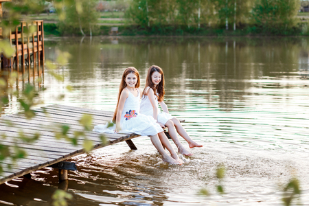 Beautiful girls friends with long brunette hair and blue eyes on the pier near the water at the sunsetの写真素材