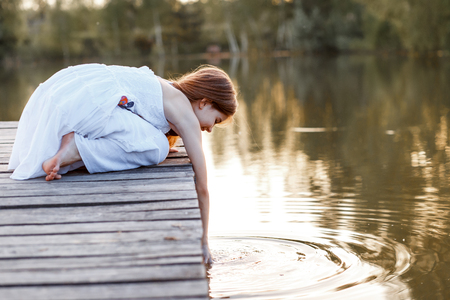 A beautiful girl with long brunette hair and blue eyes on the pier near the water at the sunsetの写真素材
