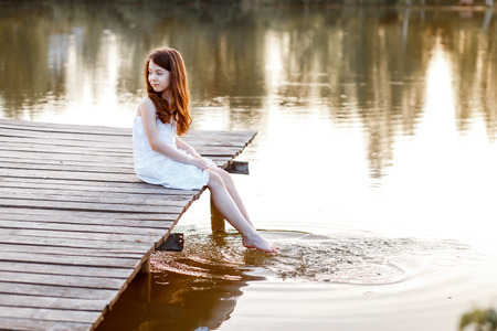 A beautiful girl with long brunette hair and blue eyes on the pier near the water at the sunsetの写真素材