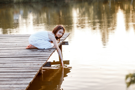 A beautiful girl with long brunette hair and blue eyes on the pier near the water at the sunsetの写真素材