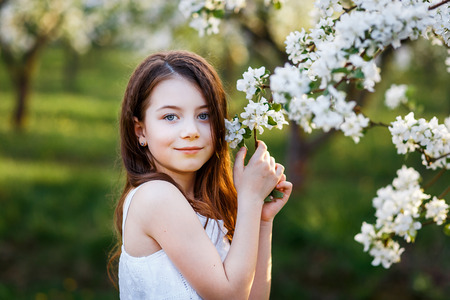 portrait of a beautiful young girl with blue eyes in white dress in the garden with apple trees blosoming at the sunsetの写真素材