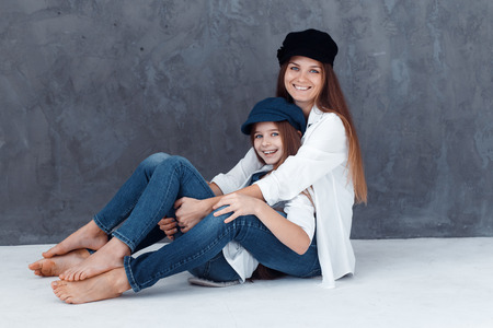 Stylish beautiful mother and a cute daughter with blue eyes in studio hugging and having fun. mom and daughter wearing hatsの写真素材
