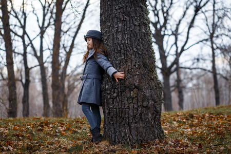 A cute little girl in grey coat and gray hat near the thick old oak in the oak wood at the sunsetの写真素材