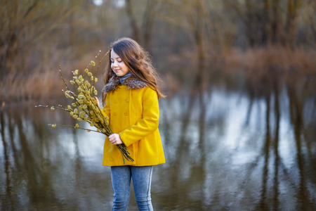 A girl in a yellow coat in the forest in early spring with a willow branch of twigs. A girl furing freshet or high waterの写真素材