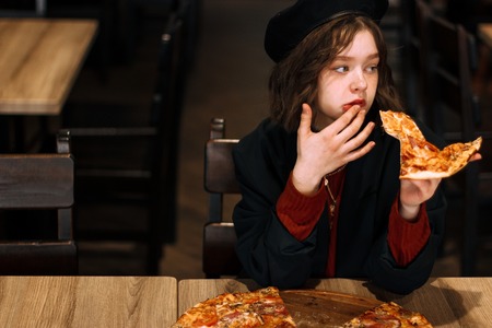 Beautiful young curly girl in black hat with piece of pizzaの写真素材