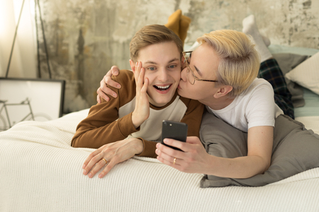 International love. Homosexual asian man with blonde hair kissing his european boyfriend and taking selfie photoの写真素材