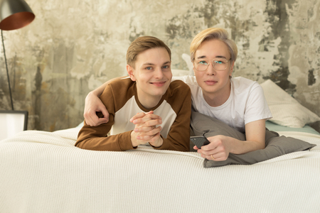 Attractive international homosexual couple relaxing in bed before night sleep and looking at cameraの写真素材