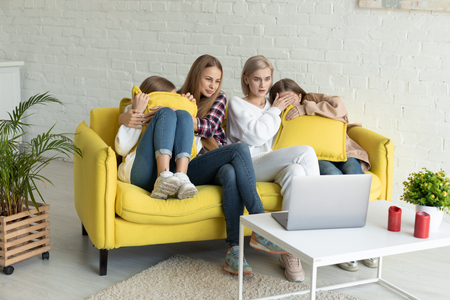 Happy young lesbian couple with daughters in casual clothes sitting together on yellow sofa at home, exited family watching a movie on a laptopの写真素材