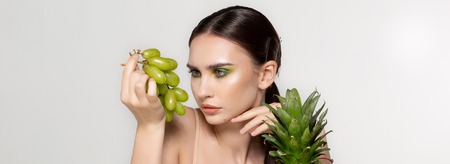 Healthy young brunette woman looking at green grapes in her hand, fruits and vegetables on the table, studio photo on grey backgroundの写真素材