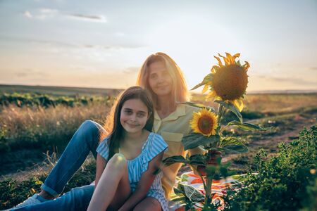 Beautiful teen girl in blue dress sitting on the plaid among the mealow near sunflowers with her young blonde mom in yellow shirt, sunset on the backgroundの写真素材