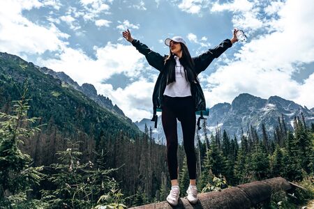 Full length portrait of young girl traveler in white hat and t-shirt that standing with open hands in front of the mountains, holding bottle of waterの写真素材