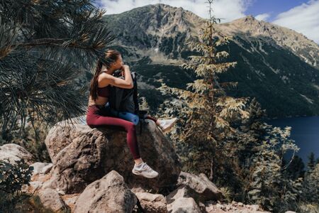 Young attractive girl in red sportswear sitting on the big stone, leaned on her backpack, tired while climbing a mountain, beautiful mountain lake on the background, active lifestyle conceptの写真素材