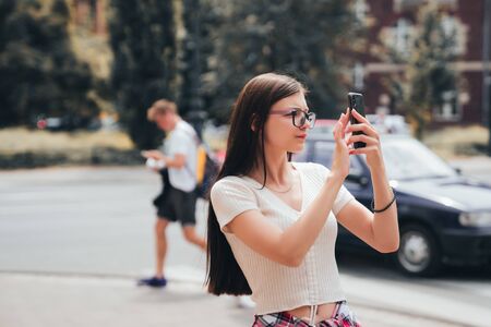 Young serious and concentrated girl in glasses standing among the streets of the city and looking for something on the phoneの写真素材
