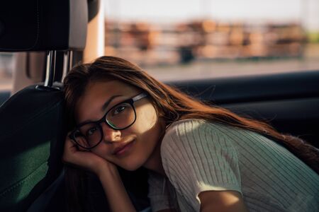 Beautiful young brunette girl in glasses sitting in the back seat of the car, leaned on her hand and...