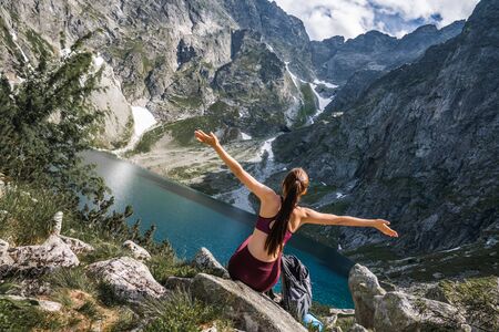 Young woman traveler in red sportswear sitting with raised hands back to the camera on the stone, beautiful mountain lake on the backgroundの写真素材