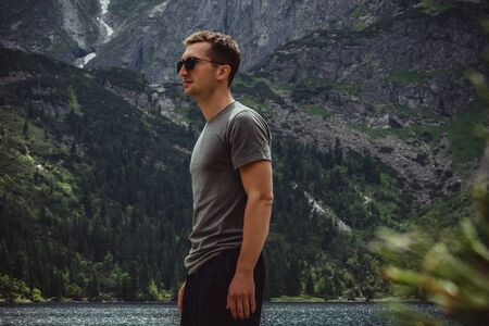 Side view portrait of young man traveler in black sunglasses and grey t-shirt in front of the mountain lake and range, active life conceptの写真素材