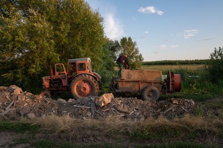 Ukraine, Chernivtsi, 10 08 2019 Old red tractor with tank standing near the lake, man in work clothes fills the barrel sprayer with waterのeditorial素材