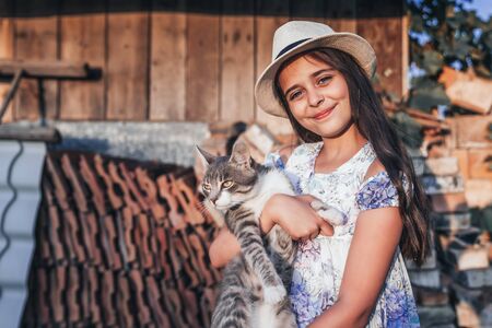 Happy smiling young girl in white hat and dress with flowers print holding a white and grey cat on her hands, looking at the camera, wooden wall and old bricks on the backgroundの写真素材