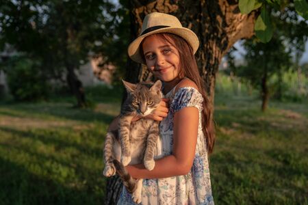 Beautiful smiling little girl in white hat holding grey cat on her hands, looking at the camera, enjoying time in the gardenの写真素材