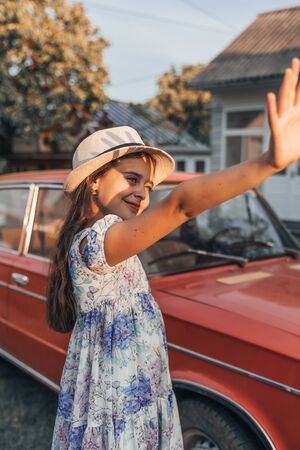 Little cute smiling girl in dress with flowers and white hat standing near red retro car and...