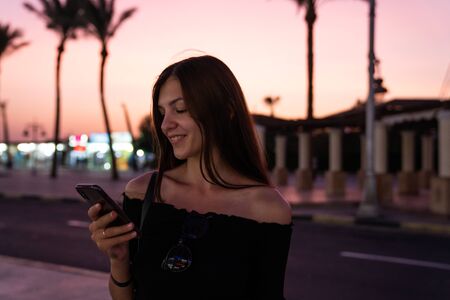 Happy brunette girl in black top reading message on her phone, blurred street with palms on the...