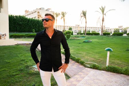 Young man in sunglasses wearing elegant clothes holding hands on his belt and looking away, sunset...