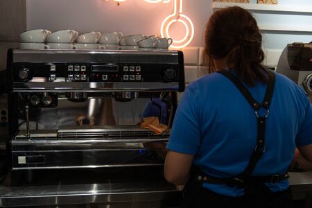 Chernivtsi, Ukraine, 25.08.2019: Brunette barista girl in blue t-shirt stands near a coffee machine back to the camera and makes a latteのeditorial素材