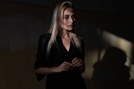 Elegant stylish young blonde woman with loose hair, dressed in black dress, stands in the shade, the rays of the sun fall on her faceの写真素材