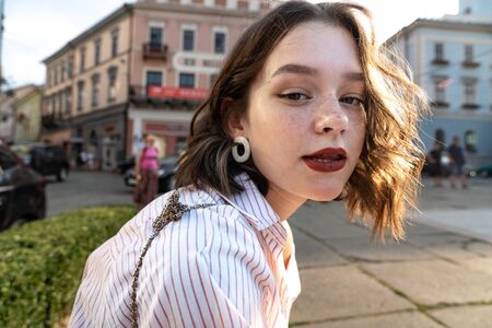 Close up portrait of the stylish serious young girl with red lips, freckles and wavy hair among the...