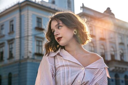 Mysterious young girl with wavy hair and red lips dressed in white striped shirt looking at the left...