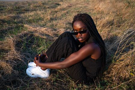 African girl in black clothes and sunglasses sits down among the dry grass field, hugging her knees...