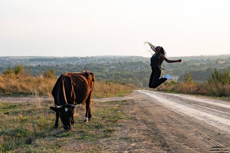 Happy african girl in black clothes jump on a dirt road, cow graze near the girlの写真素材
