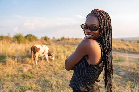 Happy smiling african girl in black sunglasses stands among the field and looking at the camera over her shoulder, cow graze on the backgroundの写真素材