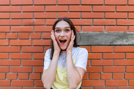 Close up portrait of the young brunette surprised girl that holds her hands on her cheeks and looking at the camera with opened mouth, isolated over red brick wallの写真素材