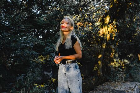 Beautiful blonde girl in black top stands among the green bushes in the shadow