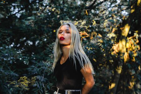 Waist up portrait of the trendy blonde girl in black top stands in the shadow among the green bushes