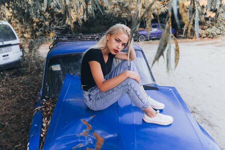 Street style fashion portrait of the blonde girl in black top and blue jeans that sits on the...