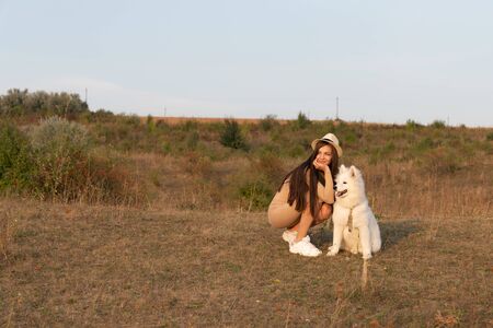 Young brunette girl in beige dress and hat crouched near the samoyed puppy, both looking at left sideの写真素材
