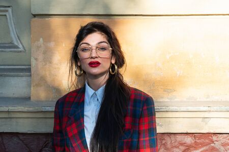 Beautiful mysterious girl brunette with glasses dressed in a plaid jacket and white shirt looks directly into the camera, the rays of the sun fall on her faceの写真素材