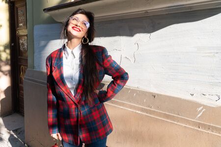 Happy girl with glasses stands in the sun and smiles, dressed in a plaid jacket and white shirt, business lady on a walk during lunchの写真素材