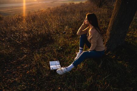 Attractive young girl in casual clothes sitting under big tree with book and looking at the beautiful sunsetの写真素材
